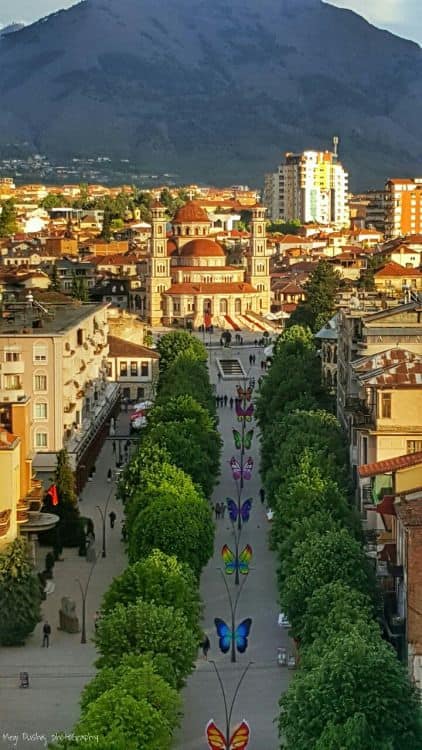 Colorful butterflies on a pedestrian street in Tirana, Albania with historic buildings and mountain backdrop.