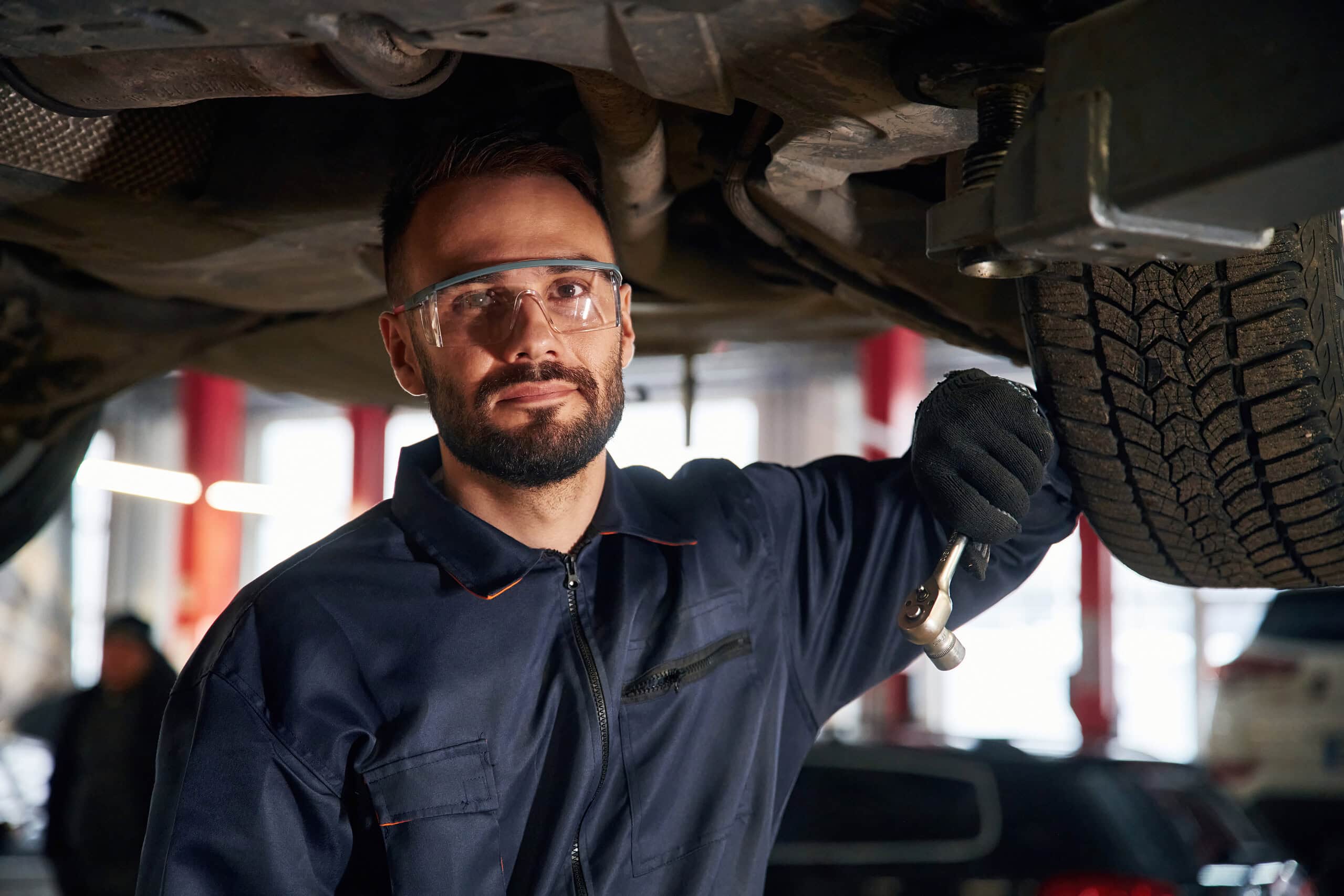 Mekanik automobilistik që kontrollon poshtë automjetit, riparim gomash, shërbim garazhi, mirëmbajtje automjetesh, riparim makinash.