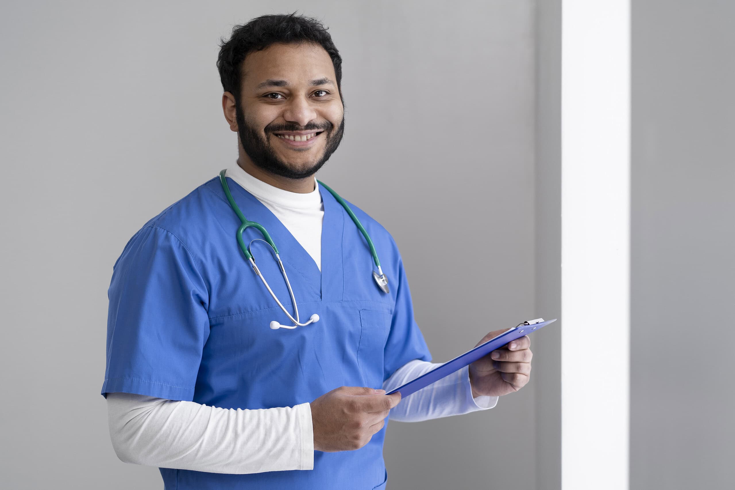 Infermiere indiane me uniforma blu duke mbajtur një tabelë shënimesh, duke buzëqeshur, në një mjedis klinik.