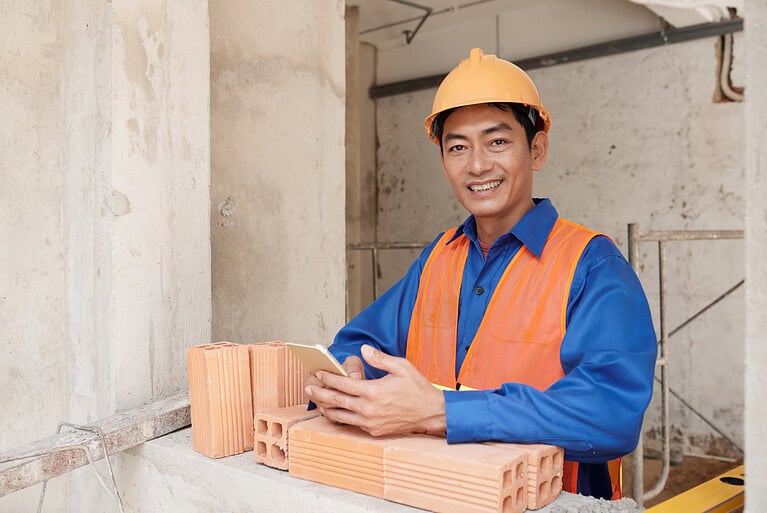 Construction worker with a safety helmet and orange vest at a building site, holding a smartphone.